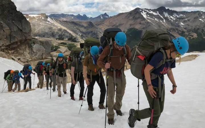 A group of people carrying backpacks and trekking poles, and wearing helmets, hike in a line up a snowy incline. Behind them is a vast mountainous landscape.